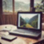 Laptop displaying a mountain scene sits on a wooden table with a notebook and charger. Window view shows more mountains. Cozy ambiance.