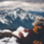 Person in winter clothing sketches in a notebook on a snowy mountain peak. Majestic snow-covered mountains fill the background under cloudy skies.