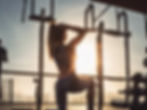 Woman in workout gear performing a pull-up in a gym during sunset. Sunlight highlights her toned back. Mood is determined and focused.
