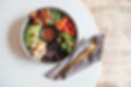 A vibrant bowl with avocados, roasted tomatoes, tofu, peas, broccoli, and sauce. Gold cutlery on a napkin beside it. White table setting.