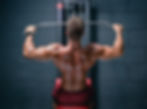 Man doing lat pulldowns in a gym, showcasing muscular back. Gray wall and equipment with some text. Wearing maroon shorts, focused mood.