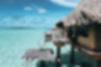 Overwater bungalow with thatched roof on turquoise sea, clear sky, distant mountains. Wooden deck and ladder leading into water. Serene vibe.