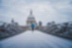 Person in blue jacket runs across a metallic bridge towards a domed building under a cloudy sky, creating an energetic and serene mood.